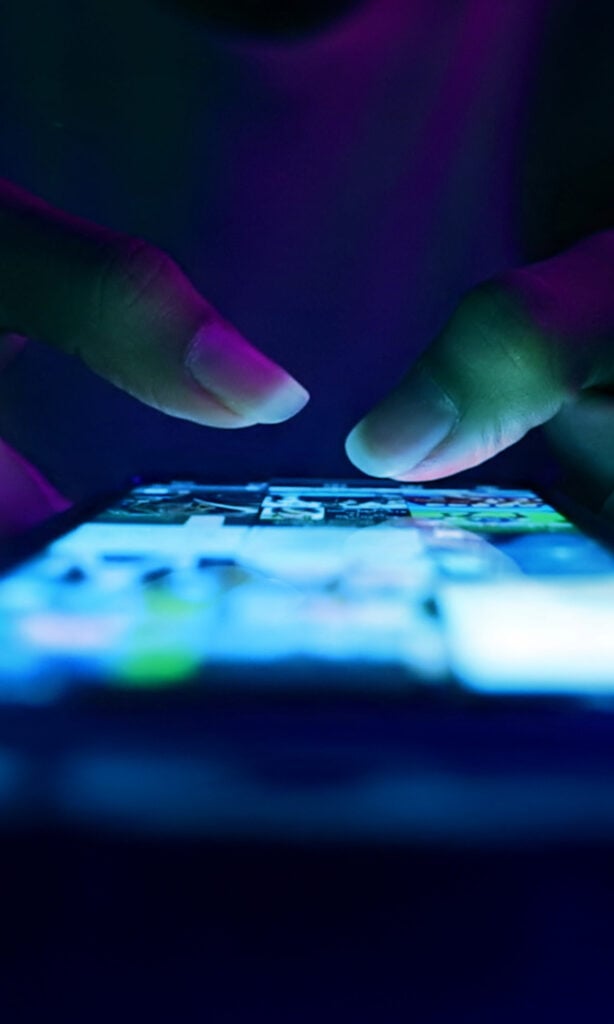 Close-up of hands using a mobile phone in a dark setting, with fingers tapping the glowing touchscreen displaying various blurred app icons, illuminated by blue and purple light.