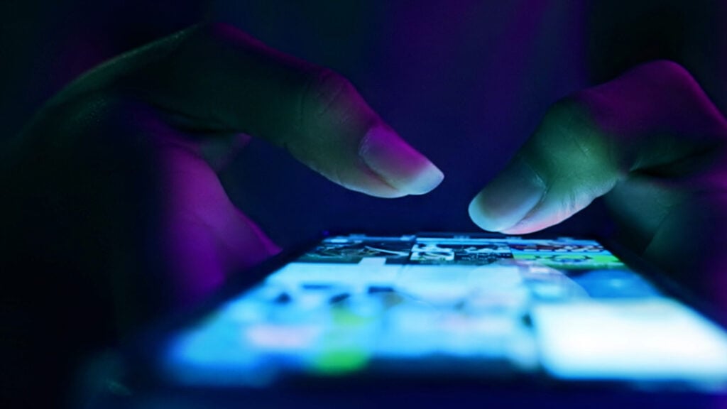 Close-up of two hands using a mobile phone in a dark setting, with the screen brightly lit and displaying various images or apps.