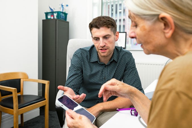 A man and an older woman sit at a desk in an office, looking at a mobile together. They appear to be discussing details as the WCRF launches CPAW, with the man gesturing while the woman holds the phone.