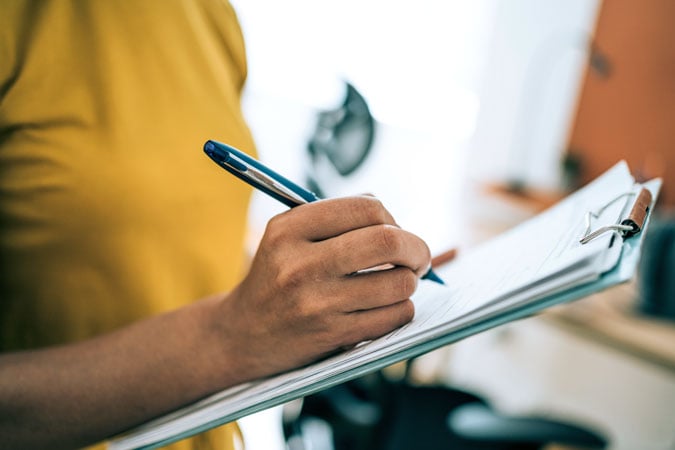 A person in a yellow shirt holds a clipboard and writes with a blue pen. The background is out of focus, suggesting an indoor setting during Cancer Prevention Action Week (CPAW) 2026.