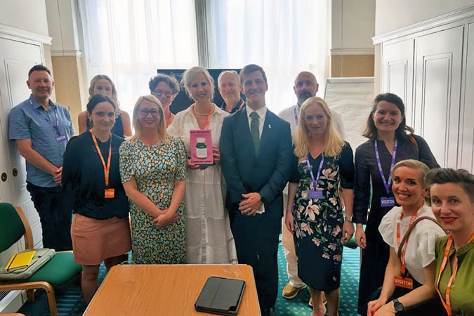 A group of thirteen people stand and smile in a well-lit meeting room. The woman in the centre holds a book on the immune system and cancer. Several people wear conference badges, and there’s a table with a tablet and notepads in the foreground.