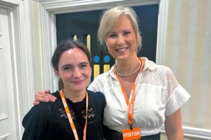 Two women, both wearing orange visitor badges, stand indoors and smile at the camera. One has her arm round the other as they pose in front of a door with a decorative design.