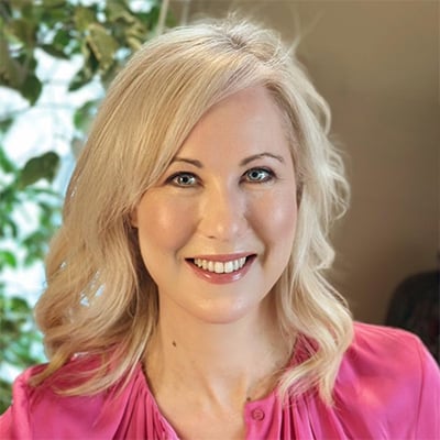 A woman with wavy blonde hair, wearing a bright pink blouse, smiles at the camera. There is a leafy green plant and a softly lit background behind her, reflecting her passion for debunking cancer diet misinformation on social media.