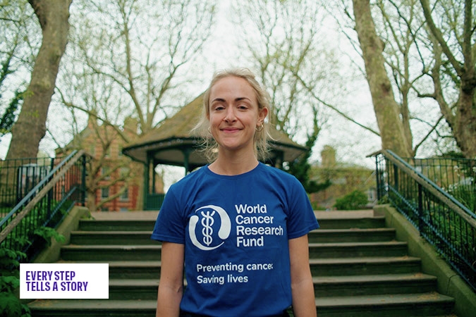 A woman stands outdoors on steps, smiling and wearing a blue World Cancer Research Fund T-shirt. Trees and a gazebo are in the background. Text in the corner reads, Every Step Tells a Story.
