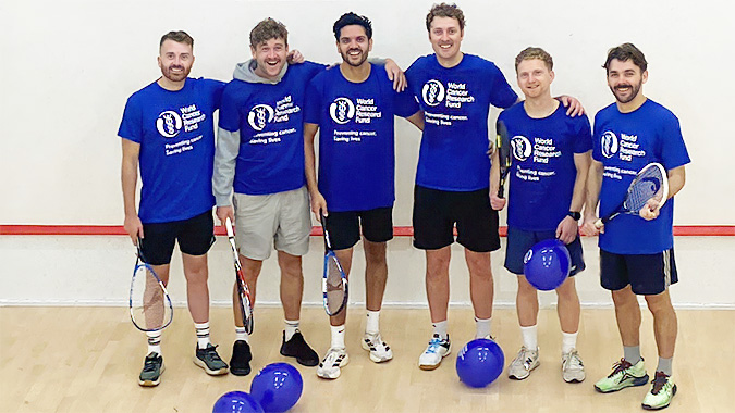 Six men in sportswear stand on a squash court holding racquets, with four blue balloons on the floor. Four of them wear matching blue shirts with a World Cancer Research Fund logo and text. They are smiling and posing together.