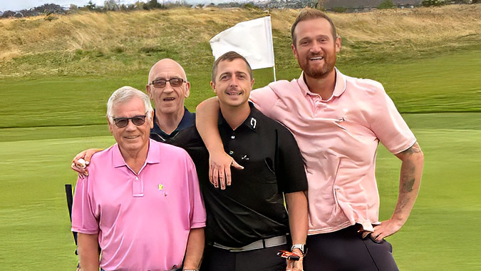 Four men stand together on a golf course, smiling at the camera. They are dressed in golf attire, with one holding golf clubs. A white flag and grassy hills are visible in the background.