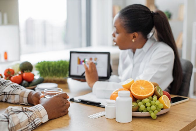 A nutritionist explains healthy eating to a client, using a tablet to dispel misinformation. Fresh fruit and vegetables, as well as supplement bottles, are on the table between them.