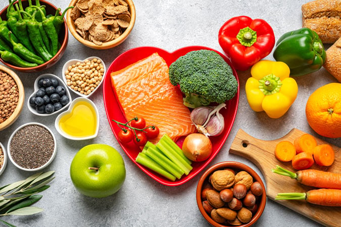 A variety of healthy foods, ideal for a cancer diet and treatment, including salmon, broccoli, peppers, fruit, nuts, seeds, grains, and olive oil are arranged on a table with a heart-shaped plate in the centre.