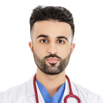 A young male doctor with short dark hair and a trimmed beard, wearing a white coat, blue scrubs, and a stethoscope around his neck, stands ready to address cancer diet misinformation on social media against a plain white background.