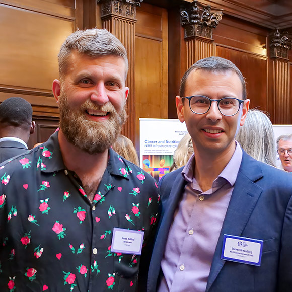 Two men stand smiling at an indoor event with wooden panelling. Both wear name badges; one has a floral shirt and a beard, the other wears glasses and a blue suit. Several people and informational posters are in the background.