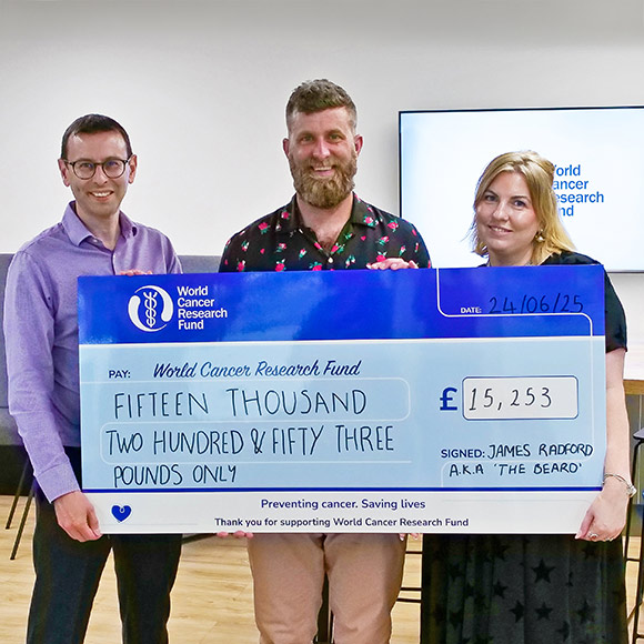 Three people stand indoors holding a large ceremonial cheque for £15,253, made out to the World Cancer Research Fund. The cheque is signed by James Radford a.k.a. The Beard.
