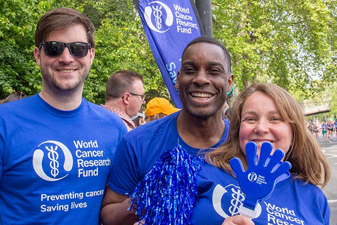 Three people wearing blue World Cancer Research Fund shirts smile at an outdoor event. One person holds a blue foam hand, and they stand in front of a WCRF banner with trees in the background.