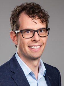 A man with curly brown hair and glasses, wearing a navy blazer and light blue shirt, smiles at the camera against a plain grey background.