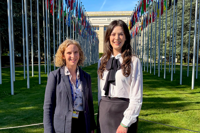 Two women stand smiling on a lawn lined with flagpoles displaying various national flags, with the United Nations building visible in the background during the World Health Assembly focussed on cancer prevention.