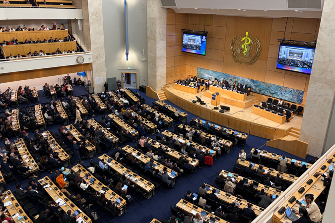 A large conference hall filled with attendees seated at rows of desks, facing a stage where speakers stand at lecterns beneath the World Health Organisation emblem; large screens display World Health Assembly cancer prevention discussions.