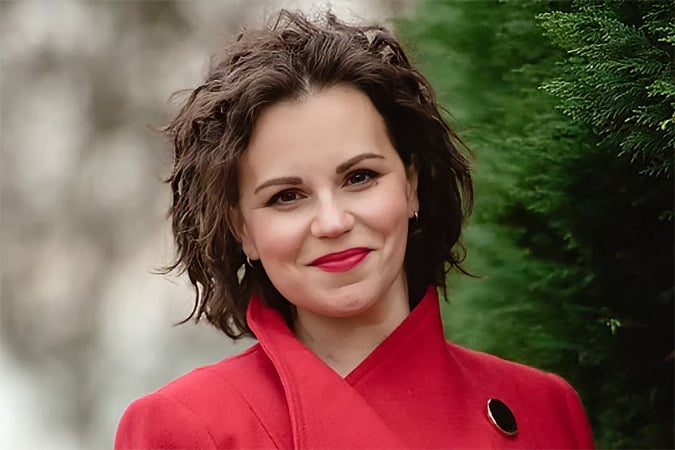 A woman with wavy brown hair, wearing a red coat and red lipstick, stands outdoors in front of green foliage and a blurred background, smiling warmly at the camera.