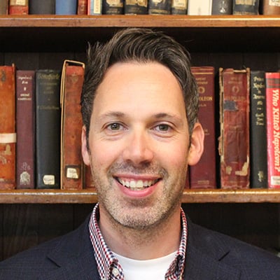 A man with short brown hair and a trimmed beard smiles at the camera, standing in front of a bookcase filled with old, colourful books. He is wearing a checked shirt and dark blazer, ready to discuss cancer diet misinformation on social media.