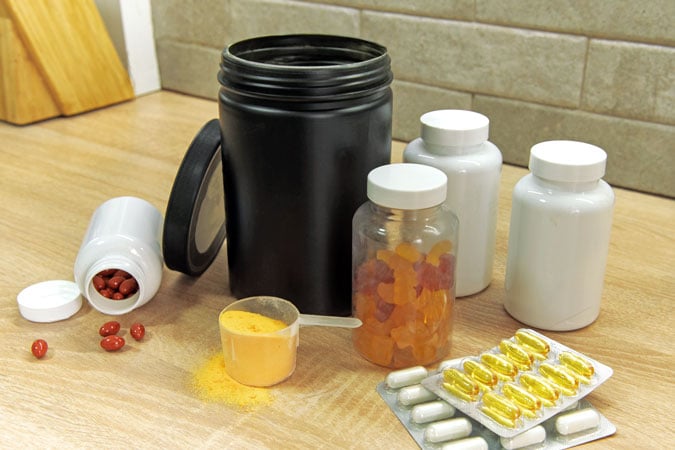 Various supplement containers on a wooden worktop, including a large black tub, white bottles, orange gummies, scattered red tablets, yellow powder with a scoop, white capsules, and cancer prevention supplements in blister packs.