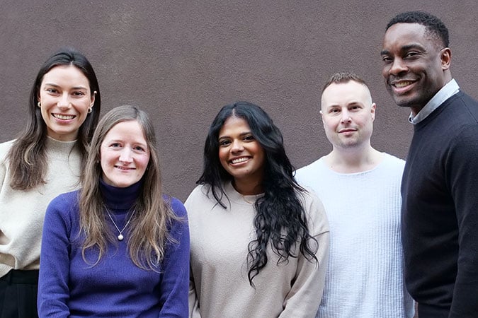 Five people standing in front of a plain brown wall, smiling at the camera. Three women and two men are wearing casual jumpers in neutral colours.