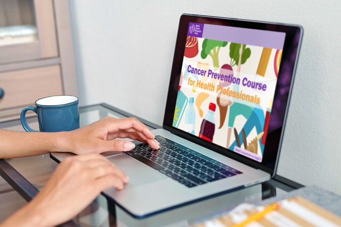A person types on a laptop displaying a colourful webpage titled Cancer Prevention Course for Health Professionals, featuring cancer prevention learning resources. A blue mug sits nearby on the glass table.