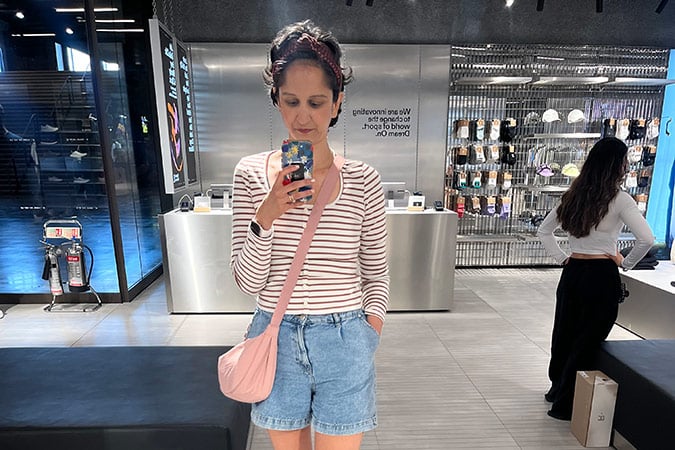 A person takes a mirror selfie in a shop, wearing a striped long-sleeved shirt and denim shorts with a pink crossbody bag, reflecting on a recent breast cancer scare.