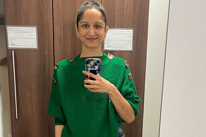 A person with short hair wearing a button-shoulder green jumper takes a mirror selfie while navigating a breast cancer scare.