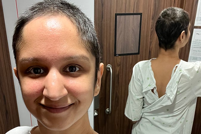 A person with a buzzcut smiles for a mirror selfie while wearing a hospital gown open at the back during a breast cancer scare.