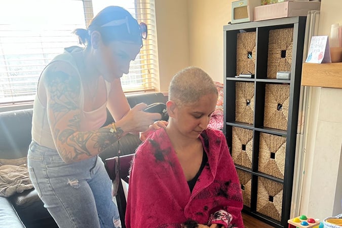 A tattooed woman uses electric clippers to shave the head of another woman draped in a pink towel during a breast cancer scare.