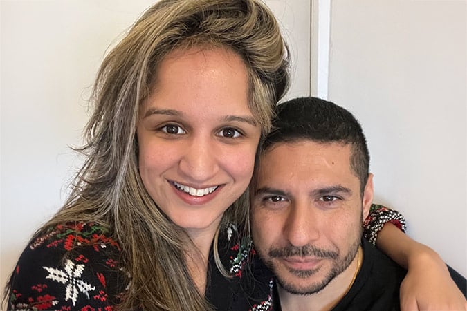 A smiling woman with long brown hair stands behind a man, arm draped over his shoulder, as they reflect on their recent breast cancer scare.