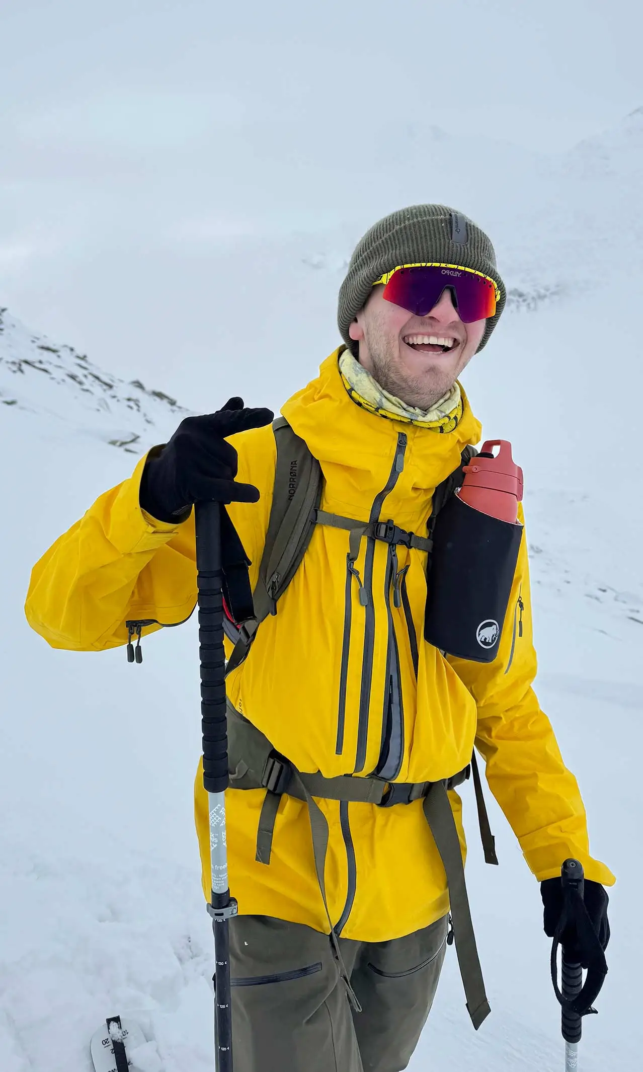 A person in a yellow jacket, green beanie, and red sunglasses smiles whilst holding a trekking pole in a snowy mountain landscape. A red water bottle hangs from their rucksack strap, proudly displaying the World Cancer Research Fund logo.
