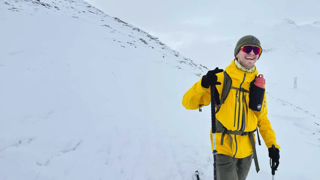 A person in a yellow jacket and sunglasses stands smiling in a snowy mountain landscape, holding ski poles and wearing a rucksack, proudly displaying support for the World Cancer Research Fund.