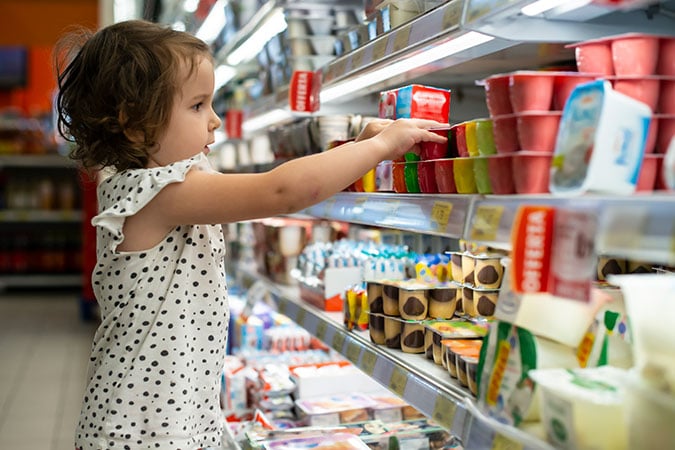 A young girl in a polka dot shirt selects yoghurt from a refrigerated shelf in a supermarket, making a choice influenced by the food environment that can impact habits related to obesity.