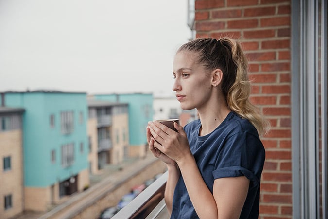 A woman with a ponytail stands on a balcony, holding a mug and looking thoughtfully into the distance. Modern apartment blocks and a street are visible in the background.