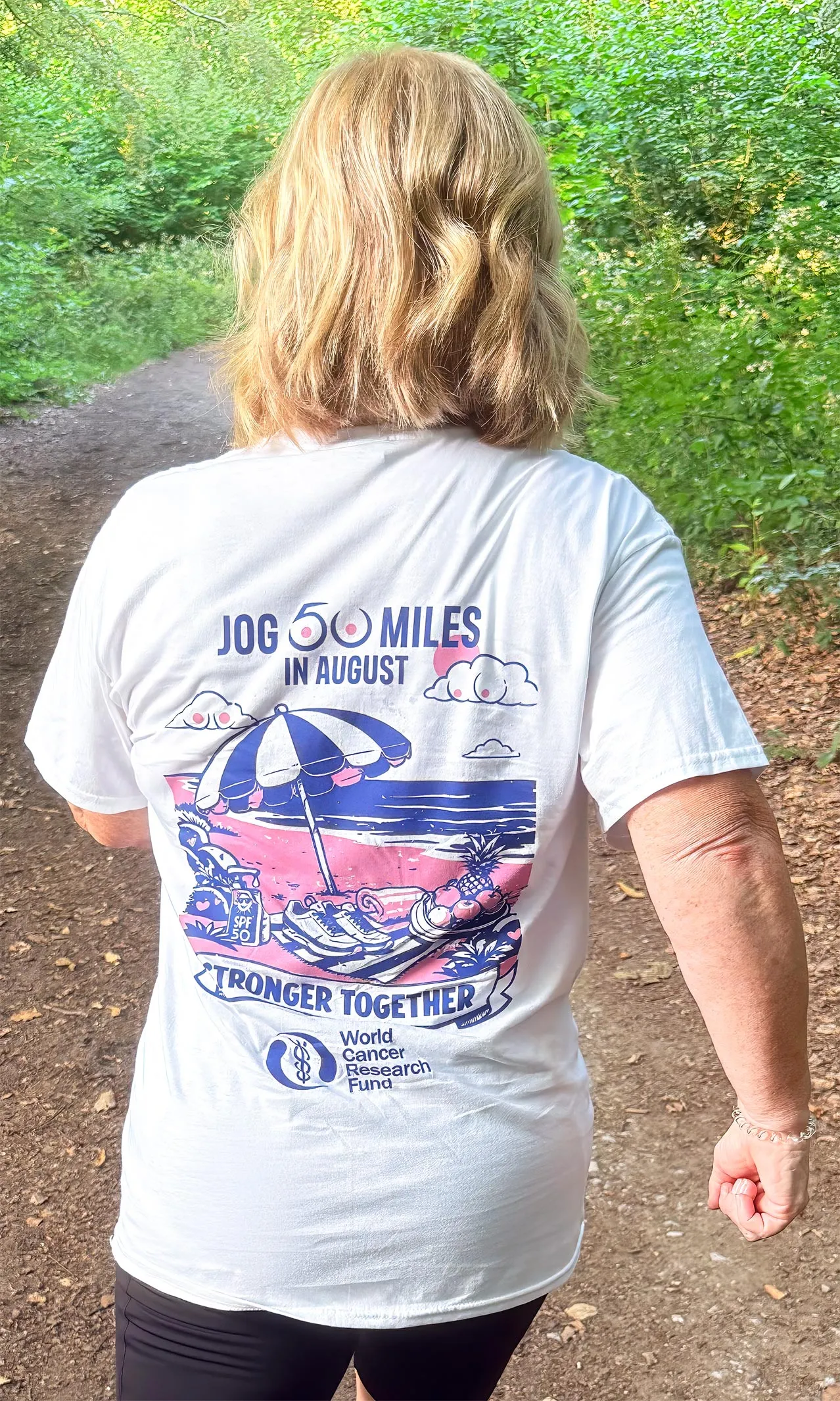 A person with blonde hair walks on a wooded path, wearing a white T-shirt that reads Jog 60 miles in August. Stronger Together. Proudly supporting the World Cancer Research Fund, the shirt features a beach-themed graphic.