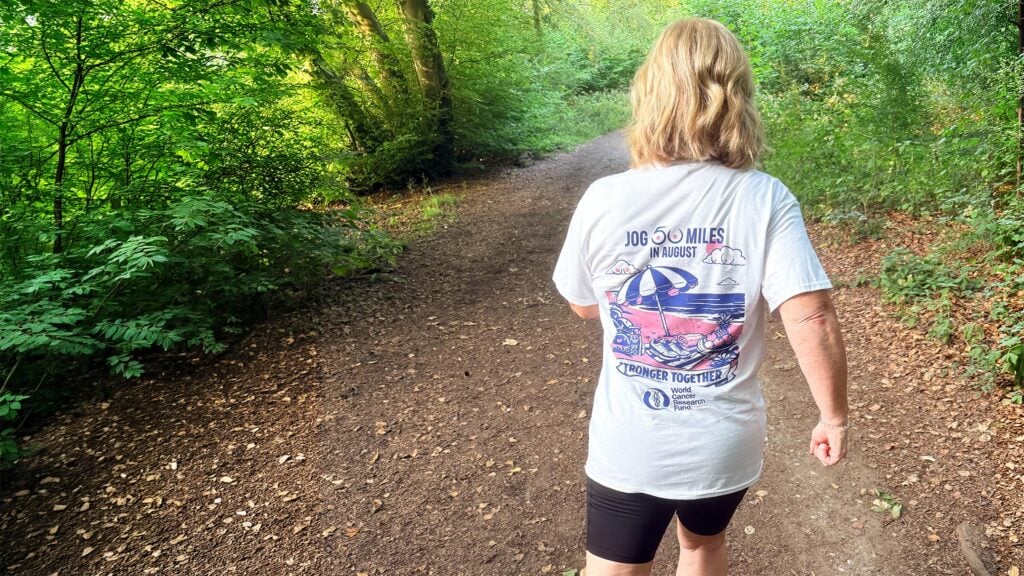 A person with blonde hair, wearing a white T-shirt and black shorts, walks on a dirt path through a green, leafy forest. The back of the T-shirt displays a colourful design and text about a 100-mile challenge.