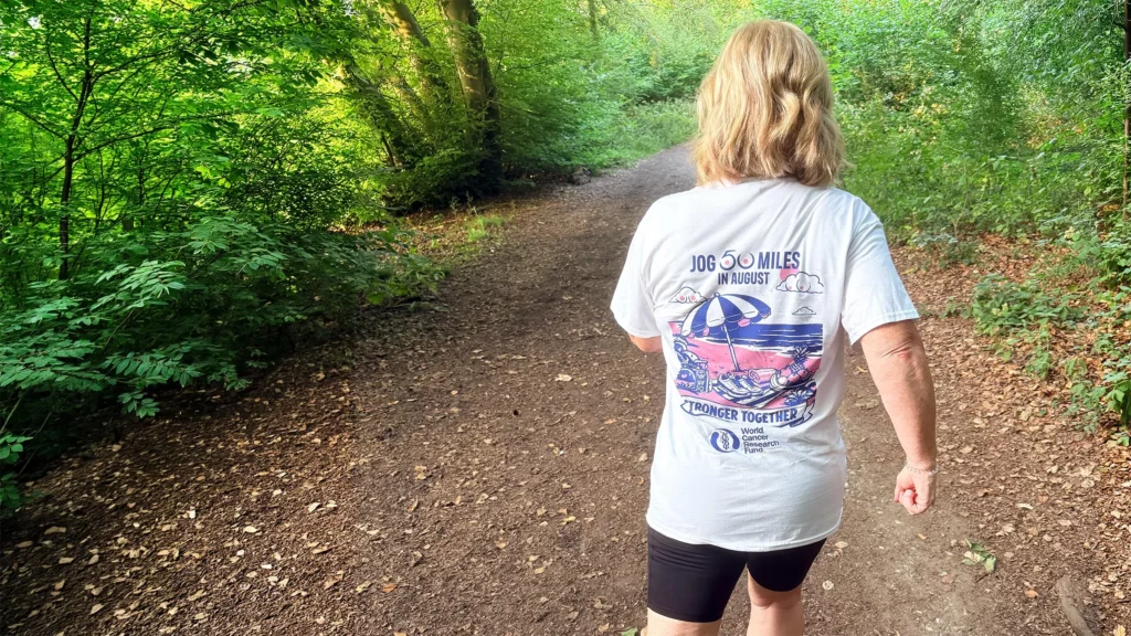 A person with blonde hair walks along a forest path, wearing a white shirt that says 100 miles in August on the back. The path is surrounded by lush green trees and sunlight filters through the leaves, as they take each step in support of the World Cancer Research Fund.