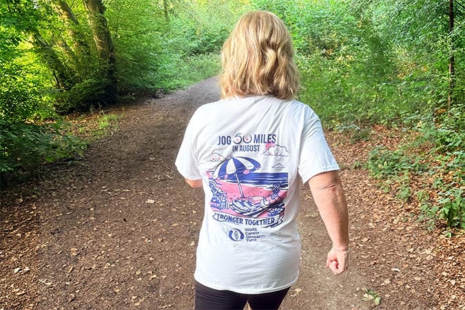 A woman with blonde hair walks on a forest trail, wearing a white T-shirt that says “Jog 50 Miles challenge in August” with an illustrated design, while trees create a scenic background.
