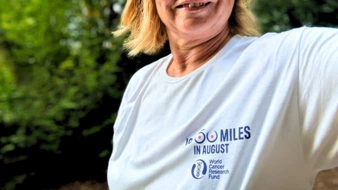 A person wearing a white T-shirt that says 60 miles in August and World Cancer Research Fund stands outdoors, smiling, with green trees in the background.