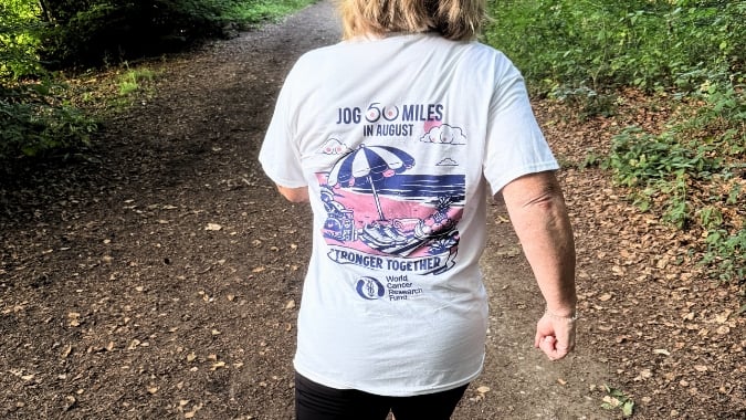 A person walks on a forest path, wearing a white T-shirt that reads Jog 50 Miles in August, Stronger Together with an illustrated beach scene and World Cancer Research Fund logo on the back.