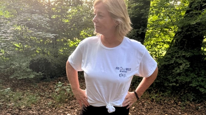 A woman with short blonde hair stands in a sunlit, leafy wood, wearing a white T-shirt with writing and black trousers. Her hands are on her hips, and she is looking to the side.