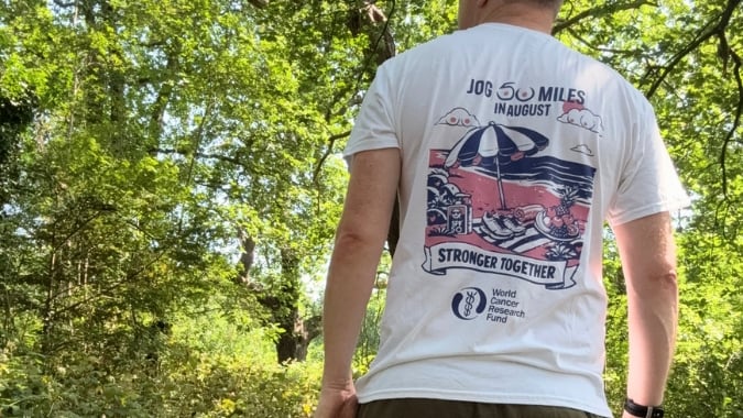 A person stands outdoors in a green, leafy area, wearing a white T-shirt with a colourful design on the back that reads Jog 50 miles in August – Stronger Together – World Cancer Research Fund.