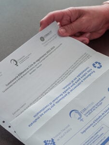 A hand holds a folded official document in Spanish with printed text and logos, including recycling and educational symbols, on a dark surface.