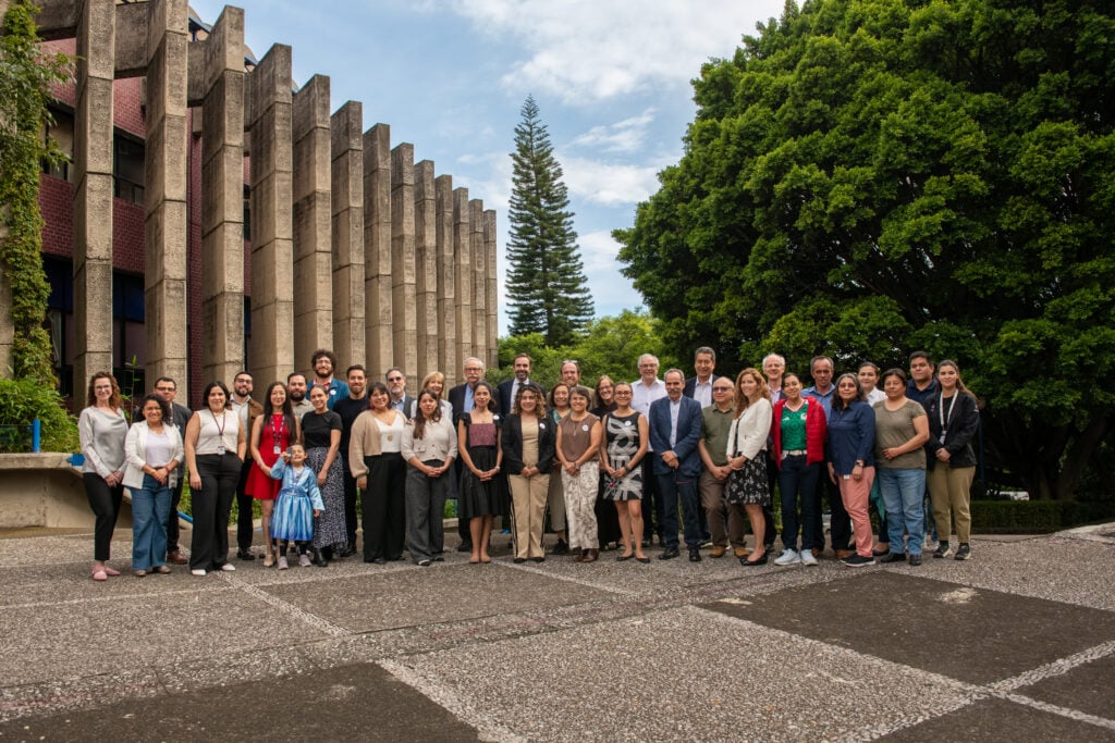 A large group of people pose for a photo outdoors in front of a modern concrete building and trees, under a partly cloudy sky.