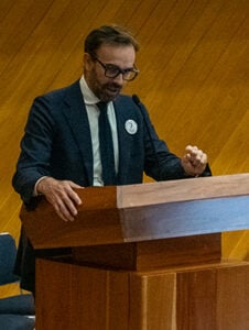 A man in a suit and glasses speaks at a wooden lectern against a wooden panelled wall.