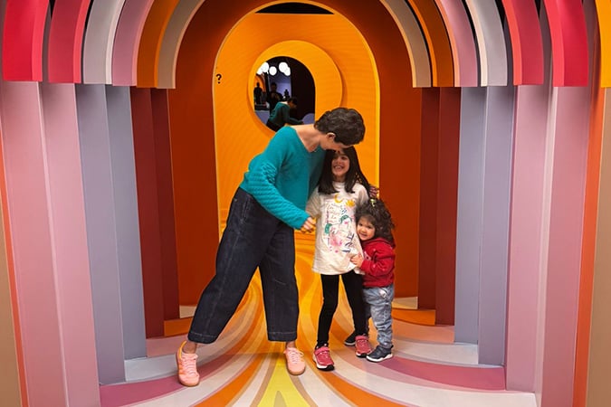 A woman and two smiling children stand together in a colourful, rainbow-like tunnel with circular arches and bright orange, pink, and yellow tones.