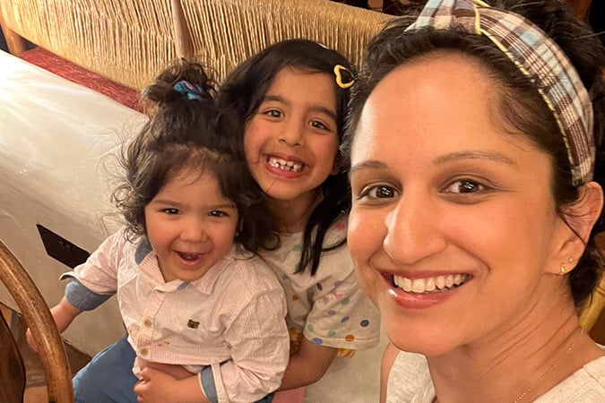 A woman smiles with two young children, one sitting on her lap and the other standing behind. They appear happy and close together indoors.