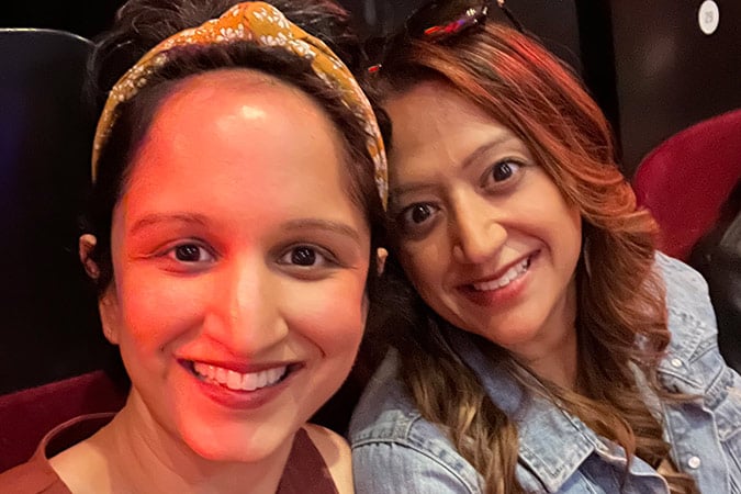 Two women smiling and sitting together in a theatre or auditorium with red and black seats. One has a yellow hairband, the other wears a denim jacket. Warm lighting shines on their faces.