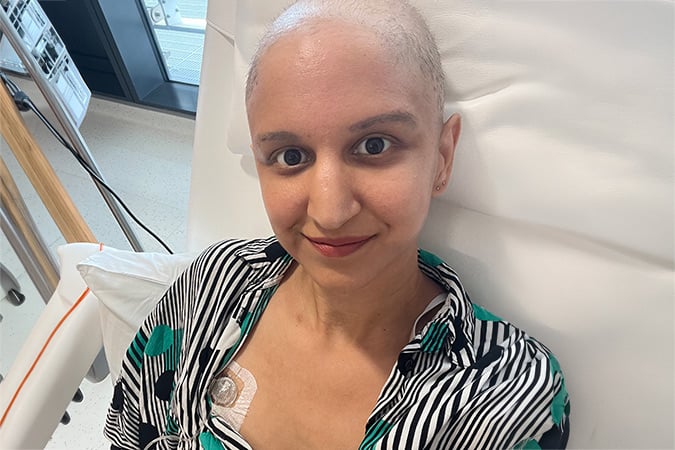 A person with a bald head sits in a hospital chair, smiling. They have a medical port visible on their chest and are wearing a striped shirt with green accents. Hospital equipment is visible in the background.