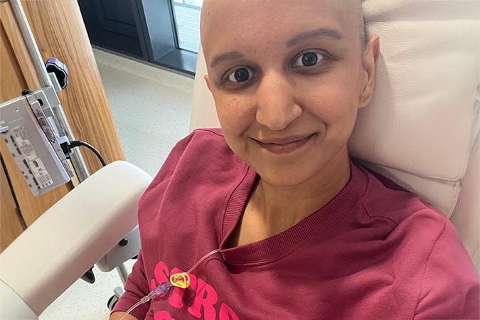 A person with a shaved head, wearing a pink shirt, smiles whilst sitting in a medical chair receiving a drip infusion. The setting appears to be a hospital or clinic.