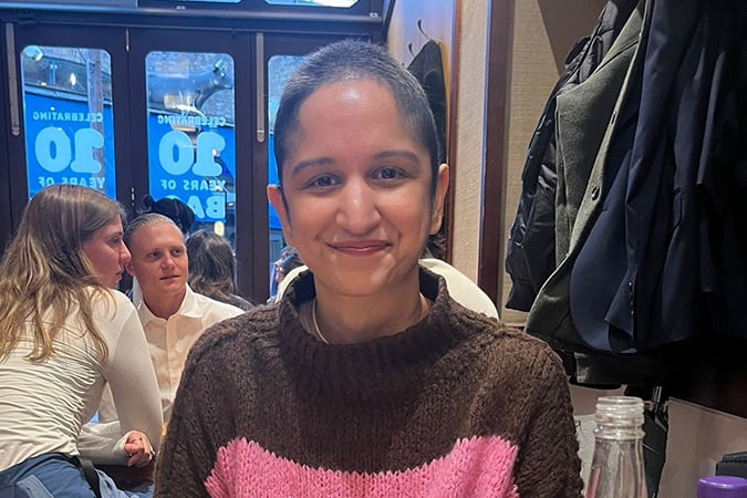 A person with short hair smiles at a restaurant table, wearing a brown and pink jumper. Jackets hang on the wall behind, and other diners are seated in the background. A water bottle is visible in the foreground.